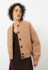 Veste marron texturée et matelassée avec un col arrondi et des boutons foncés, portée sur une robe sombre, avec un design court et des poignets élastiques.