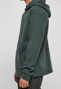 Felpa verde a costine con cappuccio e tasca frontale; vestibilità comoda e polsini a coste, abbinata a jeans scuri.