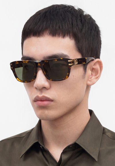 Uomo con capelli corti e scuri che indossa occhiali da sole scuri con montatura tartarugata e una camicia verde oliva con colletto su sfondo semplice.