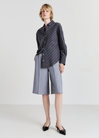 Femme debout portant une chemise à rayures foncées à boutons, un short gris clair au genou et des escarpins à talons ouverts pointus noirs.