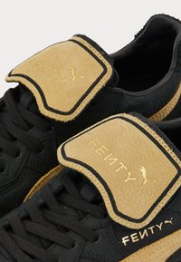 Black sneakers with a textured leather upper, featuring a tan strap with gold "FENTY" branding and black accents along the sole.