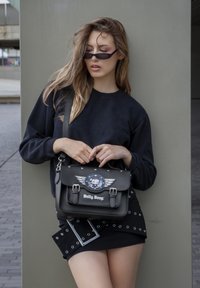 Bolso de satchel de cuero sintético negro con un gráfico circular de Betty Boop, acentos de alas, cierres de hebilla y una correa para el hombro, posicionado frente a un modelo.