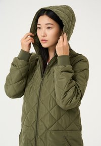 Veste matelassée vert olive avec capuche, dotée de poignets côtelés et d'une fermeture éclair frontale, tissu texturé et poches latérales.