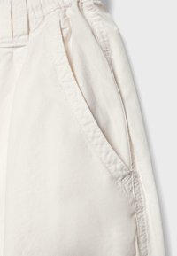 Gros plan sur une poche de pantalon beige avec des coutures visibles et une ceinture élastique sur un fond clair.