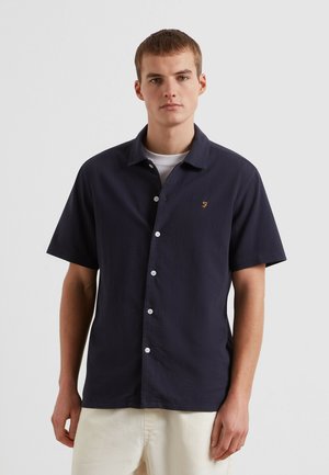 Jonge man draagt een donkerblauw kort mouw overhemd over een wit T-shirt met lichtbeige broek tegen een effen achtergrond.