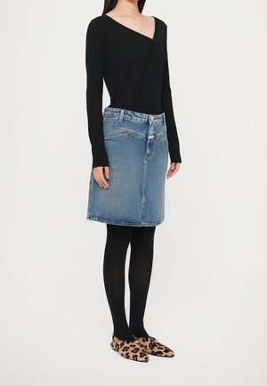 Person iført asymmetrisk sort langærmet top, blå denim nederdel, sorte strømpebukser og leopardmønstrede spidse flats stående på ensfarvet baggrund.
