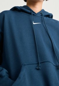 Persona vestida con una sudadera de Nike azul oscura, con bolsillo delantero y cordones, mostrando solo la parte superior del torso y el cuello contra un fondo claro.