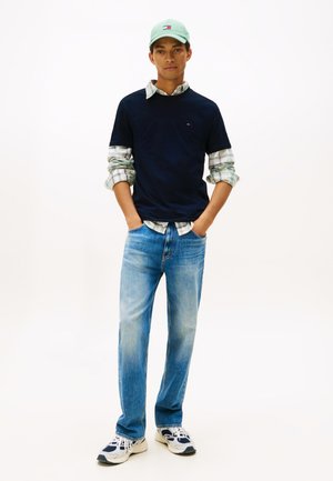 Young man stands with hands in pockets wearing a mint green cap, navy t-shirt over a plaid shirt, light blue jeans, and white sneakers.