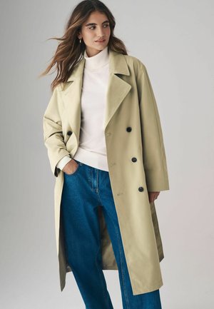 Hellbeige doppelreihiger Trenchcoat mit großen Reverskragen, dunklen Knöpfen und oversized Passform, kombiniert mit einem weißen Rollkragenpullover und blauen Jeans.