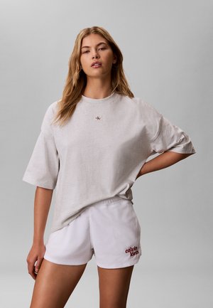 Joven mujer con camiseta oversize gris claro y pantalones cortos blancos de Calvin Klein, de pie con una mano en la cadera sobre un fondo liso.