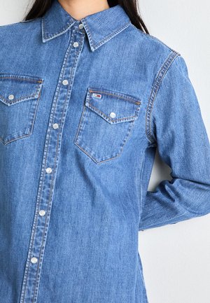 Woman wearing a blue denim button-up shirt with two chest pockets and snap buttons, standing against a white background.