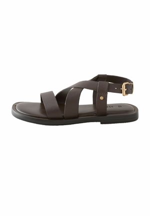 Tamsiai rudos odos lygios basutės su susikertančiomis dirželiais ir reguliuojamu auksiniu sagtimi ant balto fono.