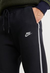 Crne dukserice od pamuka, s uskim fitom, sivim naglascima uz strane i vidljivim Nike logotipom na lijevoj nozi.