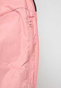 Ljusrosa, slät tygjacka med ett inre foder som har en ficka med dragkedja och kontrasterande sömdetaljer längs kanterna.