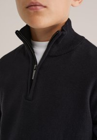 Tuvuma persona, kas valkā melnu džemperi ar rāvējslēdzēju virs baltas T-krekla ar apaļu kakla izgriezumu, rāvējslēdzējs daļēji atvērts pie kakla.