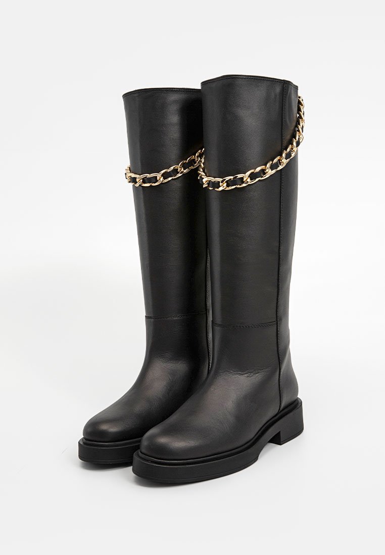 Schwarze Leder-Stiefel bis zum Knie mit einer groben Sohle, die einen goldenen Kettenakzent am oberen Schaft für zusätzliche Details aufweisen.