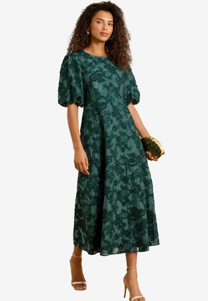 Abito midi verde ricamato con maniche a sbuffo, tessuto testurizzato e motivi floreali. Abbinato a una pochette dorata e tacchi nudi.