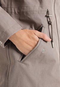 Giacca in tessuto grigio con tasca frontale, dettagli in metallo e chiusura con coulisse. Tessuto liscio con dettagli di cuciture visibili.