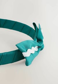Un bandeau en tissu turquoise avec une surface texturée et un accent en nœud agrémenté d'une bordure en zigzag blanche. Le design est flexible et léger.
