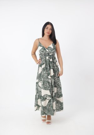 Kobieta o długich ciemnych włosach, ubrana w maxi sukienkę bez rękawów z zielonymi liściastymi wzorami i białe sandały z odkrytymi palcami, stoi z jedną ręką na biodrze.