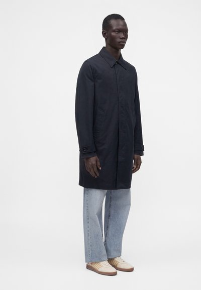 Grande personne portant un long manteau bleu marine foncé, un jean large bleu clair et des baskets blanches, se tenant devant un fond uni clair.