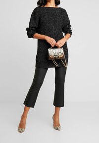 Nainen pukeutuneena mustaan neulemekkoon, mustiin lyhennettyihin nahkahousuihin, kädessään pieni käärmekuvioinen käsilaukku ja jalassa siihen sopivat käärmekuvioiset korkokengät.