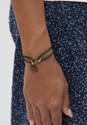 Groene koordarmband met een gouden schakelhanger en een ronde bedel met de inscriptie "CPM STUDIOS." Textuur van donkerblauwe stof op de achtergrond.