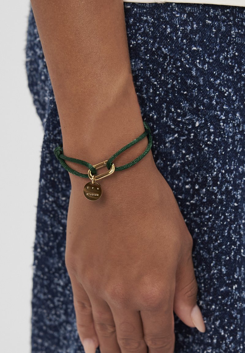 Braccialetto con corda verde con un anello in metallo dorato e un ciondolo circolare inciso con "CPM STUDIOS". Sfondo in tessuto blu navy testurizzato.