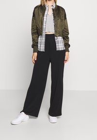 Olivově zelená saténová bomber bunda přes šedý crop top, v kombinaci s černými širokými kalhotami a bílými teniskami. Viditelná kostkovaná košile.