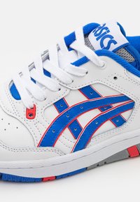 Baskets blanches Asics avec accents bleus et rouges, lacets blancs, et logo de la marque visible sur la languette rembourrée bleue et grise.