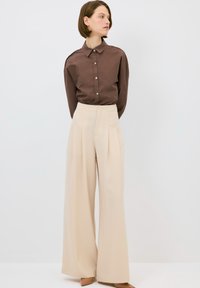 Camicia marrone con bottoni e colletto, abbinata a pantaloni beige lunghi e larghi. Tessuto morbido, con pieghe sottili sui pantaloni.