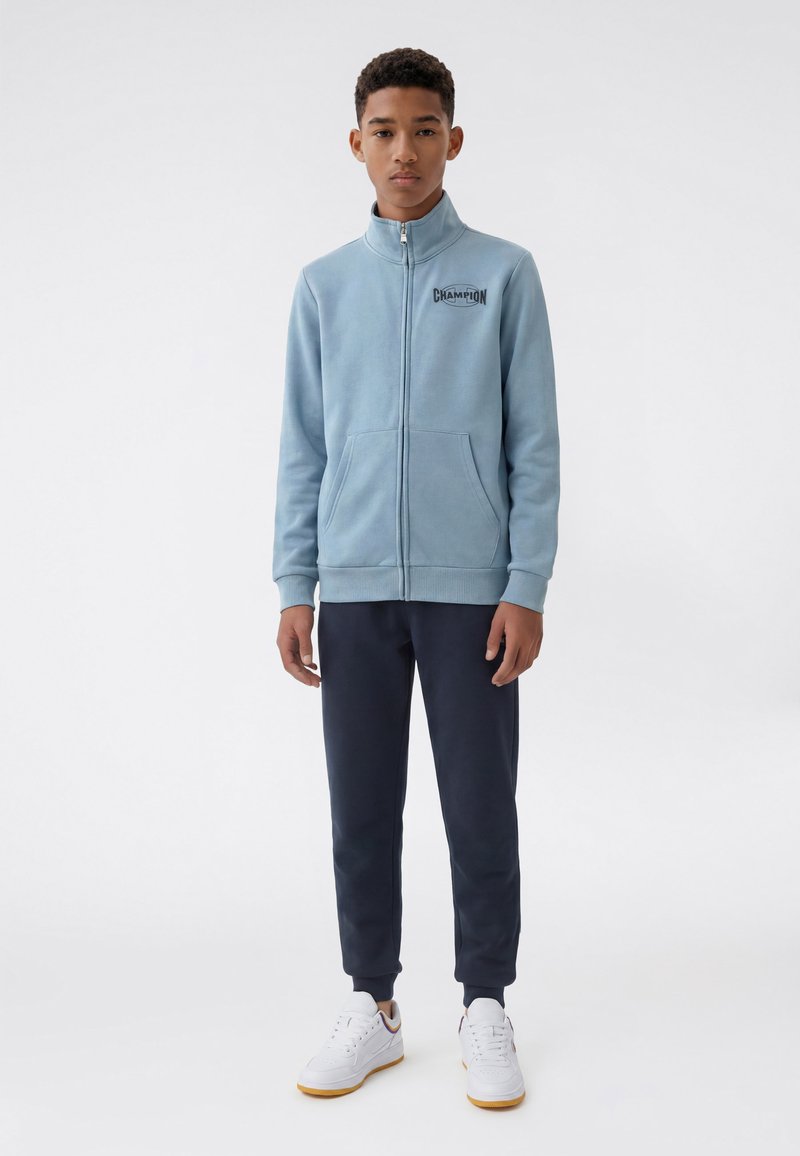Adolescent portant une veste zippée bleu clair avec logo Champion, pantalon foncé et baskets blanches, debout devant un fond uni.