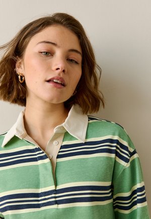 Jeune femme aux cheveux courts marron, portant des boucles d'oreilles créoles dorées et une chemise rayée verte, marine et crème à col, sur fond beige.