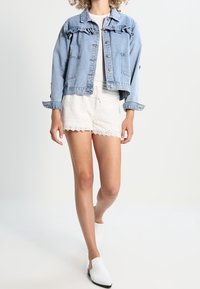 Giacca di denim con bordi arruffati, di colore azzurro chiaro. Pantaloni corti in pizzo bianco. Mules bianche con tacco basso. T-shirt semplice sotto.