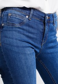 Jeans de mezclilla azul con un diseño ajustado, líneas limpias, cinco bolsillos y cierre de botón metálico. Textura suave con detalles de desvanecimiento ligero.