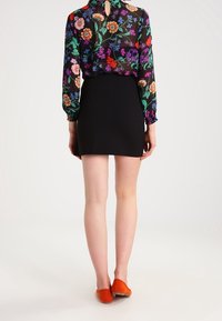 Schwarze Bluse mit floralem Muster und langen Ärmeln, Rückendetail mit Schlüssellochausschnitt, kombiniert mit einem eng geschnittenen schwarzen Rock und orangefarbenen Ballerinas.