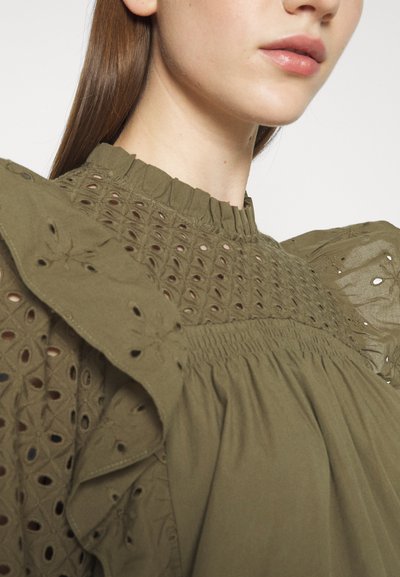 Top blouson vert olive avec des épaules à volants, comportant des découpes brodées, une taille froncée et un col montant avec un détail plissé.