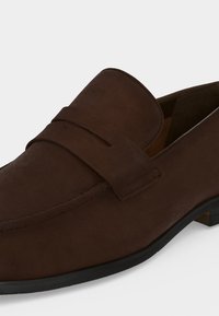 Mocassin homme en daim marron avec une bride sur le dessus et une semelle noire basse visible de profil.