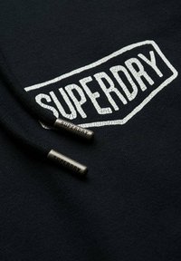 Czarna bluza z białym, pękniętym logo "SUPERDRY". Posiada dwa czarne sznurki ściągające z teksturowanymi metalowymi końcówkami, na których jest wygrawerowane "SUPERDRY".