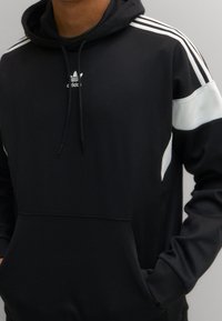 Black hooded sweatshirt with white accents and three white stripes on each shoulder. Features a front pocket and a logo on the chest.