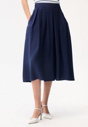 Marineblauwe midi-rok met plooien, gedragen met witte enkellaarsjes met bandje en een gestreept shirt, handen in de zakken, staand tegen een effen achtergrond.