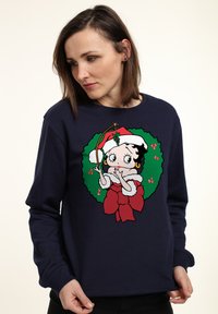Námořnická modrá mikina s vánočním motivem postavy s červenou mašlí a zeleným věncem, vyrobená z měkké tkaniny.