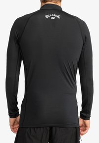 Rash guard de manga larga negro hecho de una tela suave y elástica. Presenta costuras planas y un logo en la parte superior de la espalda.