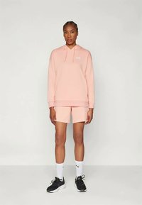 Gaiši rozā kapucīte ar priekšējo kabatu un stringiem, apvienota ar atbilstošām rozā šortiem; melni sporta apavi un baltas zeķes papildina apģērbu.