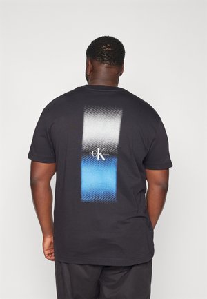 Homme portant un t-shirt noir Calvin Klein Jeans avec un design rectangulaire en dégradé blanc et bleu dans le dos, debout devant un fond uni.