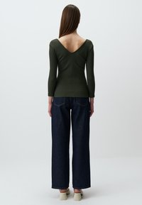 Top lungo a maniche lunghe di colore verde oliva a coste con scollo a V profondo sul retro, abbinato a jeans a gamba larga blu scuro e scarpe chiare. Tessuto morbido.