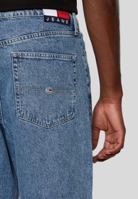 Ljusblå denimjeans med en bakficka, vertikal sömnad och en etikett i rött, vitt och blått. Texturen verkar vara slät.