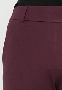 Gros plan d'une personne portant un pantalon taille haute couleur prune avec un détail passant de ceinture et un haut noir.