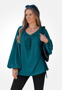 Teal gekleurde blouse met textuur, een ronde halslijn en trekkoorddetail. Voorzien van lange ballonmouwen en een losse pasvorm, gecombineerd met zwarte broek.