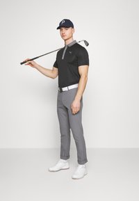 Black polo shirt with gray collar, gray tailored pants, white belt, and white golf shoes. Holds a golf club, wearing a navy hat.